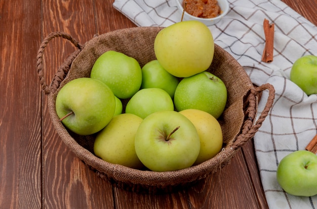 Вид сбоку зеленых и желтых яблок в корзине с яблочным вареньем и корицей на клетчатой ткани и деревянном столе