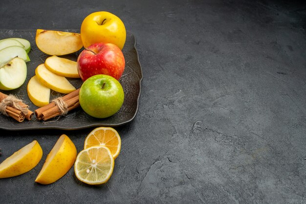 Side view of green yellow and red sliced and whole fresh apples on a black plate and cinnamon limes on the right side on a dark table