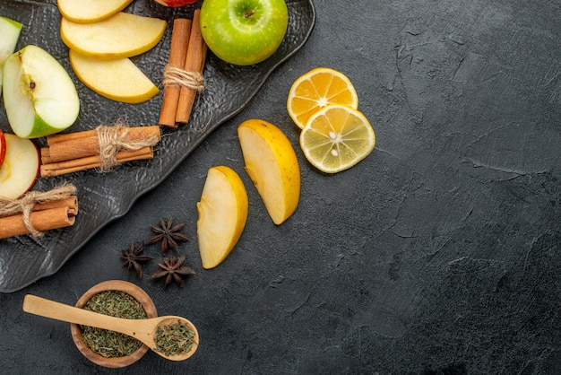 Side view of green yellow and red sliced and whole fresh apples on a black plate and cinnamon limes on the right side on a dark table