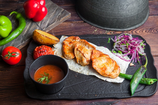 Side view grilled chicken with potatoes, tomato, onion, pepper and sauce on a dark wooden table horizontal