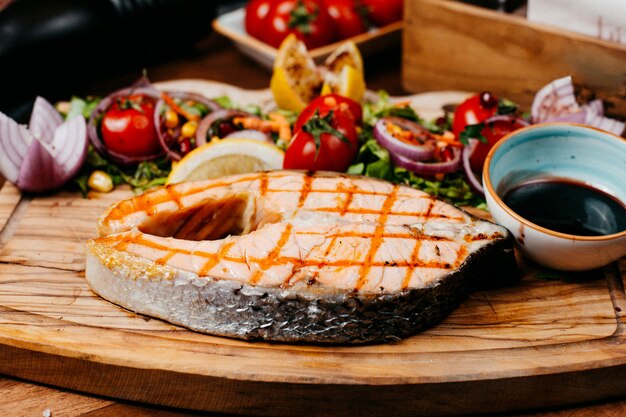 Side view of grilled salmon served with vegetables and sauce on wooden board