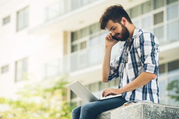 Side view of guy in casualwear answering phone call while computing on laptop outdoors