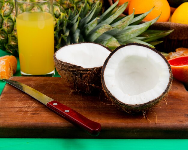 Side view of half cut coconut and knife on cutting board with other citrus fruits and orange juice on green background