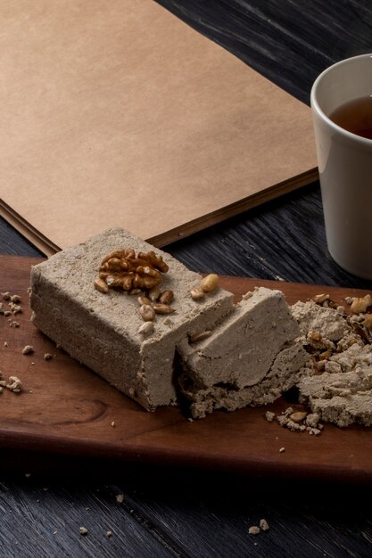 Side view of halva with sunflower seeds and walnuts on a wooden board and a cup of tea on rustic