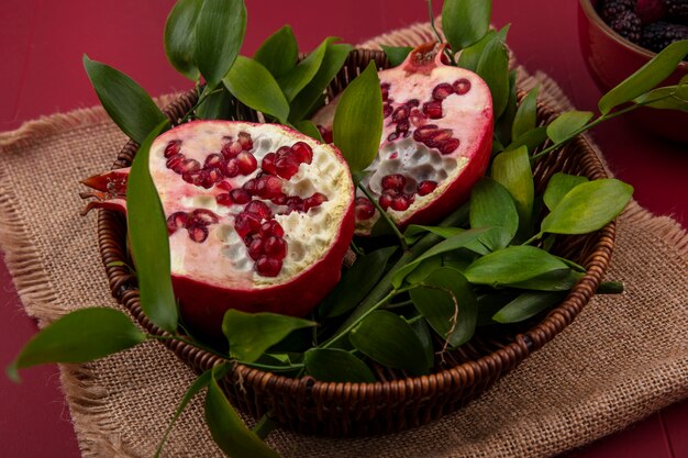 Side view of halves of pomegranate with leaf branches in a basket on a beige napkin red surface