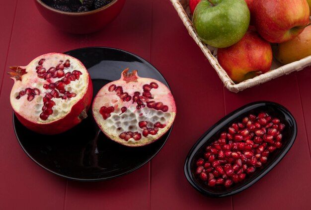 Side view of halves of pomegranates on a black plate with colored apples in a basket on a red surface