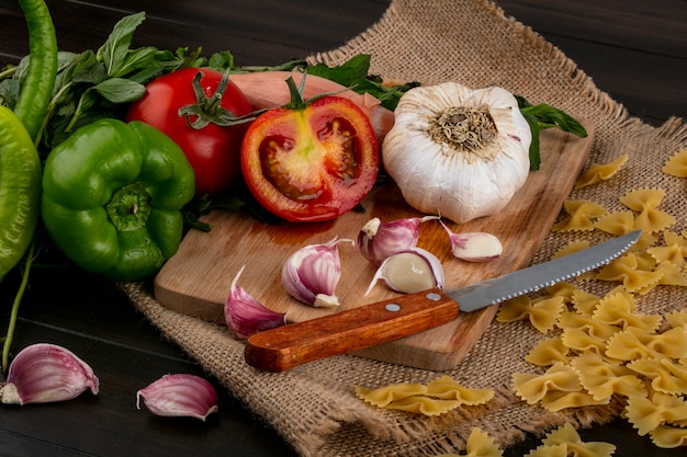 Вид сбоку половинки помидора с ножом и чесноком на разделочной доске с пучком мяты и сырых спагетти на бежевой салфетке