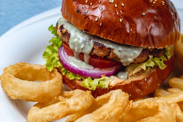 Side view hamburger grilled beef patty with melted cheese red onion tomato lettuce between burger buns and onion rings on the table