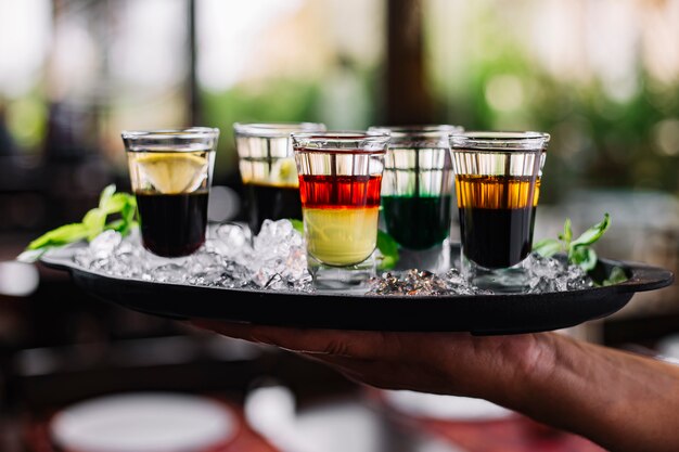 Side view of a hand holding a tray with ice and colorful cocktails in shot glasses