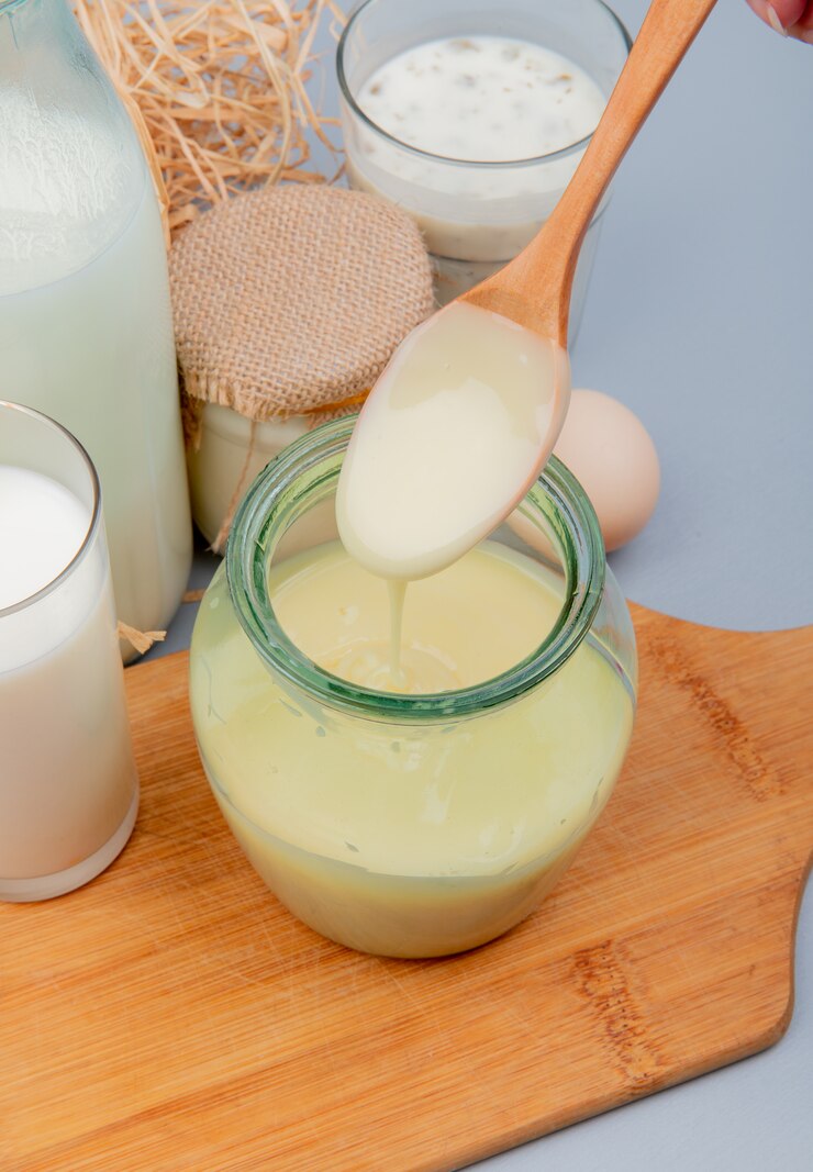 Free Photo side view of hand holding wooden spoon with condensed milk in it and glass of milk on cutting board cream egg yogurt soup on blue surface