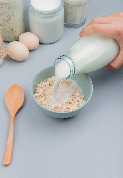 Side view of hand pouring milk in bowl with oat-flakes and spoon eggs yogurt soup sour clotted milk on blue surface