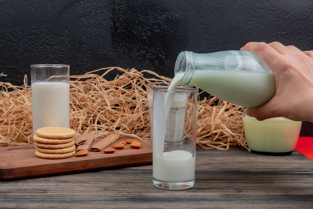 side view of hand pouring milk from bottle into glass and cookies almonds glass of milk on cutting board with yogurt soup condensed milk straw on wooden surface and black wall