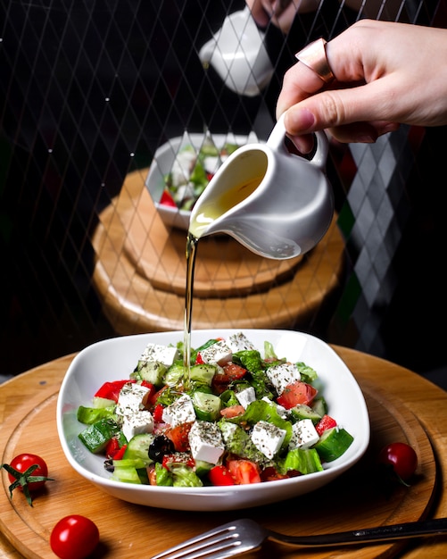 Side view of hand pouring olive oil on fresh salad with feta cheese tomatoe cucumbers in white bowl