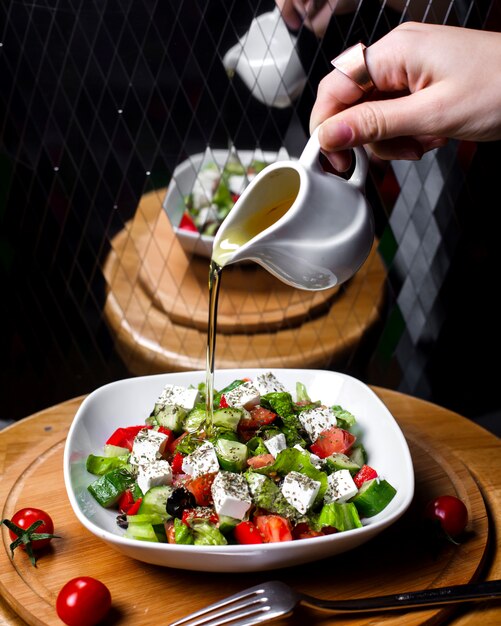 Вид сбоку руки наливая оливковое масло на свежий салат с сыром фета помидоры огурцы в белой миске