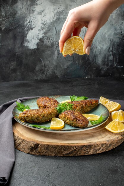 Side view of hand squeezing limon on delicious cutlets served with greens on wooden cutting board black towel on dark color background