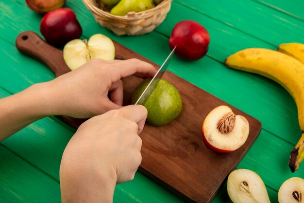 Side view of hands cutting apple with knife and half peach on cutting board with banana and half cut pear on green background