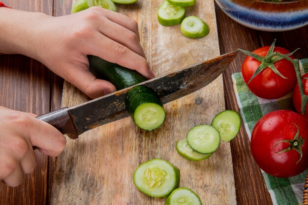 Side view of hands cutting cucumber with knife on cutting board with tomatoes on wooden surface