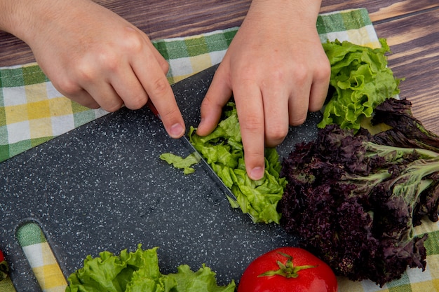 Side view of hands cutting lettuce with knife basil on cutting board and tomato on cloth and wooden surface