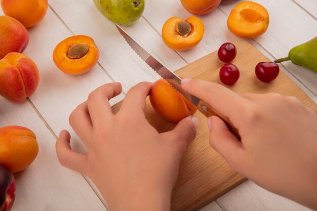 Side view of hands cutting peach with knife and cherries on cutting board with pattern of pears peaches on wooden background