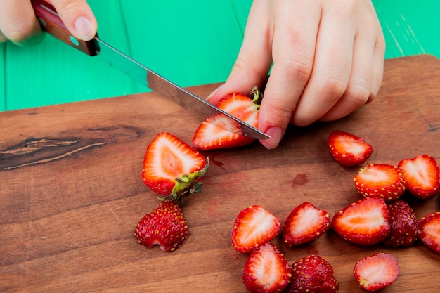 Side view of hands cutting strawberries with knife on cutting board on green surface