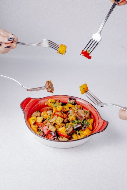 Вид сбоку руки держат вилки над миской с тушеным мясом, картофелем и овощами