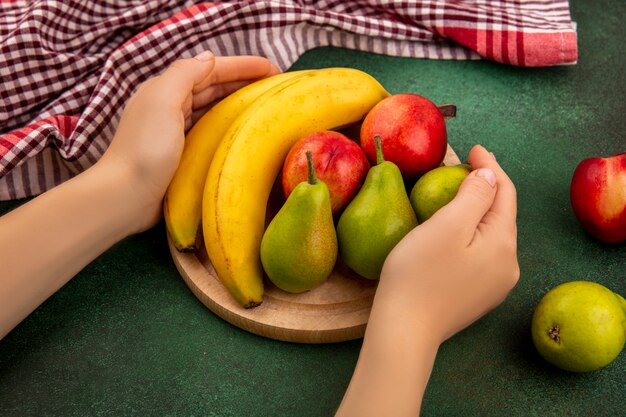 녹색 배경에 격자 무늬 천으로 복숭아 배 바나나로 그것에 과일과 함께 커팅 보드를 들고 손의 측면보기