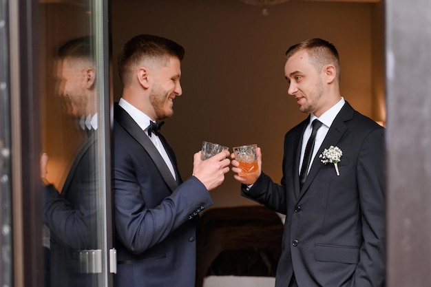Side view of handsome men in elegant suits clinking glasses celebrating wedding day having stagparty Standing in a cozy apartment and smiling Friend of the groom