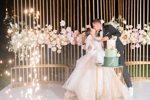 夜の式典で金属とバラで飾られた美しい結婚式のアーチの背景に立ってウエディングケーキを食べて、お互いにキスをする幸せな新郎新婦の側面図