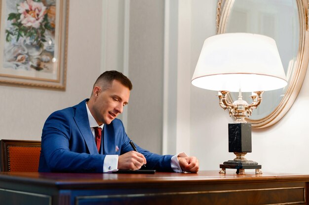 Side view of happy bridegroom wearing in stylish blue jacket white shirt and red tie concentrating on writing text on digital device while sitting at table in room with vintage decor