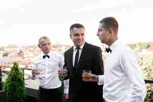 Free photo side view of happy groom in stylish black suit holding glass with alcohol and listening greets of friends while standing on terrace outdoor during sunny wedding day