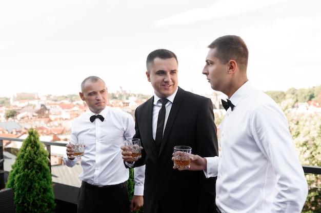 Side view of happy groom in stylish black suit holding glass with alcohol and listening greets of friends while standing on terrace outdoor during sunny wedding day