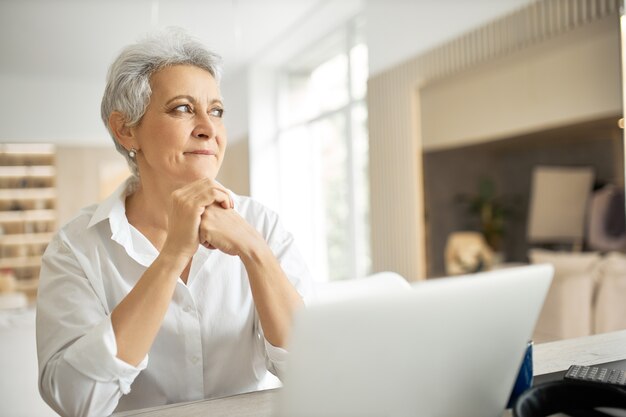 Side view of happy middle aged businesswoman with short gray hair working on laptop in her stylish office with hands on keyboard, typing letter, sharing good news