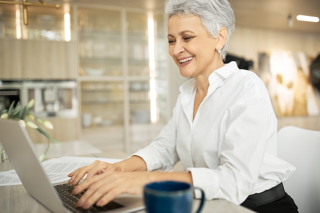 Vista laterale di felice di mezza età imprenditrice con corti capelli grigi che lavora al computer portatile nel suo elegante ufficio con le mani sulla tastiera, digitando la lettera, condividendo buone notizie