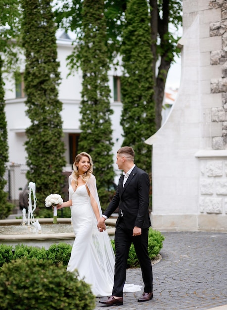 Side view of happy newlyweds wearing in wedding apparel holding hands looking to each other and enjoying atmosphere while walking in park on background of fountain architecture buildings and plants