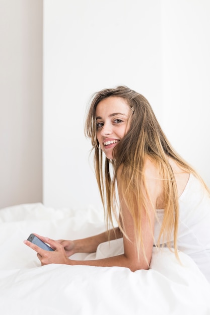 Вид сбоку счастливая женщина, сидя на кровати, держа мобильный телефон