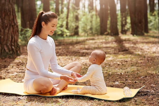 Вид сбоку счастливой молодой спортивной матери, сидящей на каремате в лесу со своим младенцем