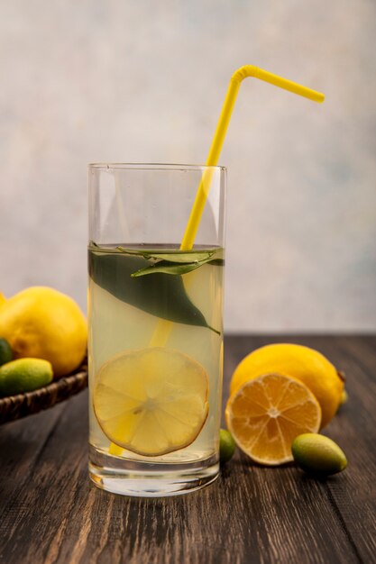 Side view of healthy lemon juice in a glass with lemons and kinkans on a wicker tray on a wooden table on a white surface