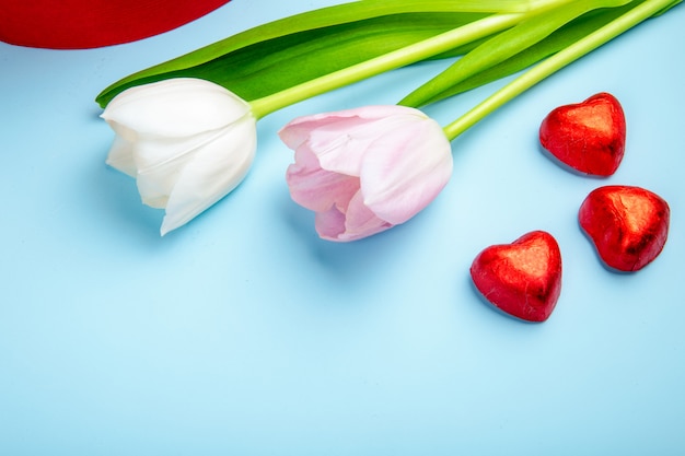 Side view of heart shaped chocolate candies in red foil with pink and white color tulips on blue table