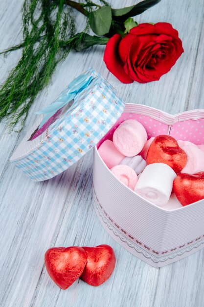 Side view of a heart shaped gift box filled with pink marshmallow and chocolate candies wrapped in red foil and red rose flower on grey wooden table