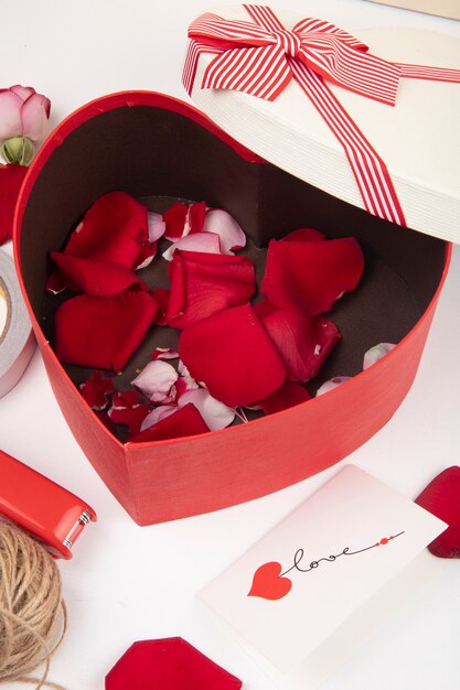 Side view of heart shaped gift box filled with red rose petals on white background