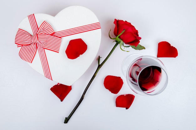 Side view of a heart shaped gift box and a glass of wine with red color rose and petals on white background