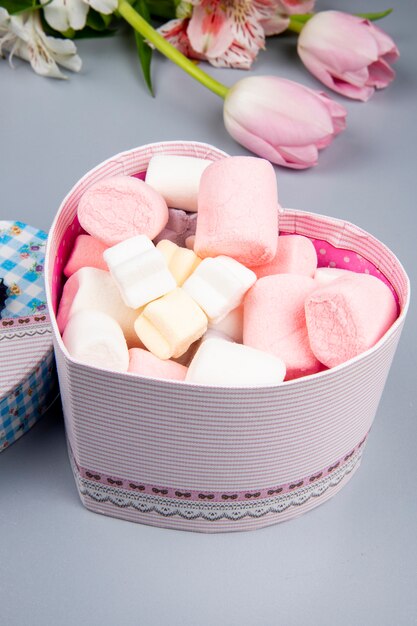 Side view of a heart shaped present box filled with marshmallow and pink tulips with alstroemeria flowers on white table