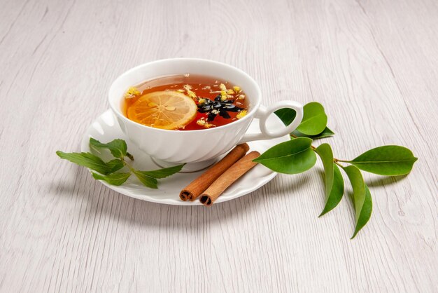 Вид сбоку травяной чай чашка травяного чая с лимоном и палочками корицы на блюдце и листьями на белом столе