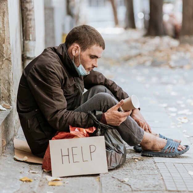 Бездомный мужчина на открытом воздухе, вид сбоку со знаком помощи и чашкой