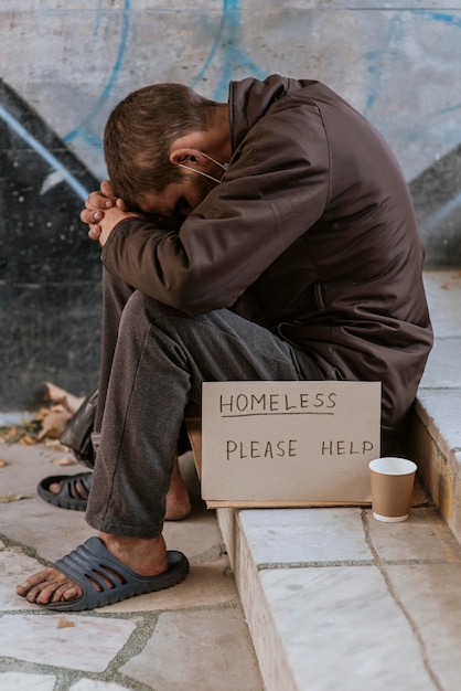 Side view of homeless man on stairs with cup and help sign