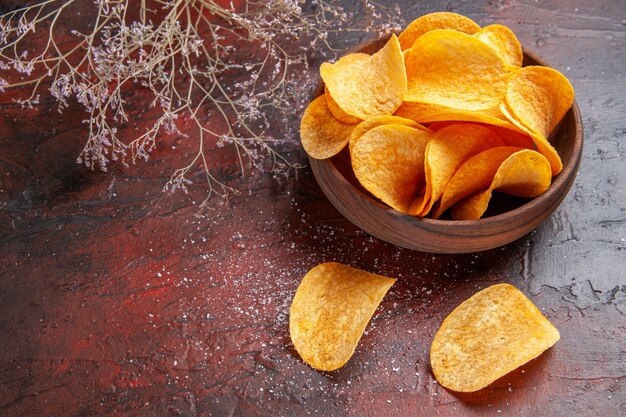 Side view of homemade delicious crispy potato chips inside and outside of brown pot on dark background