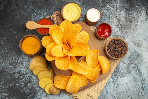 Вид сбоку домашних вкусных картофельных чипсов на деревянной разделочной доске, различные специи и майонезный кетчуп на газете на сером столе