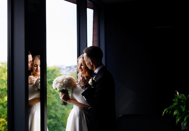 Side view of incognito bride groom in trendy black suit with modern watch on hand kissing girl face and embracing her shoulder while