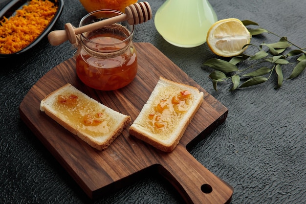 Side view of jar with quince jam and bread slices smeared with quince jam on cutting board and grated orange zest jar of lime juice half cut lemon and leaves on black background