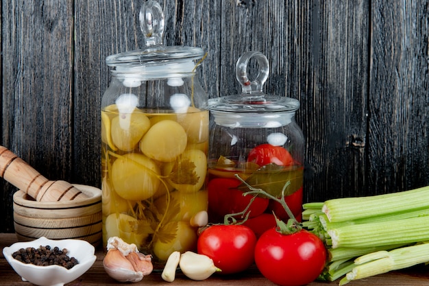 Free photo side view of jars of pickled tomato with vegetables black pepper and garlic crusher on wooden background with copy space